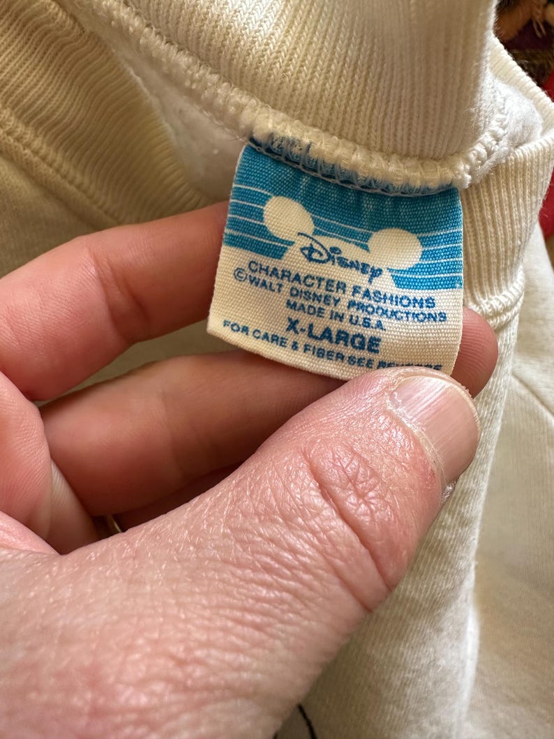 Op de afbeelding: Close-up van een blauw en wit Disney-label op een cr&egrave;mekleurig kledingstuk. Het label leest "CHARACTER FASHIONS" en "MADE IN U.S.A." met het Disney-logo en de tekst "X-LARGE". Het kledingstuk is gemaakt van een zachte, geribbelde stof.