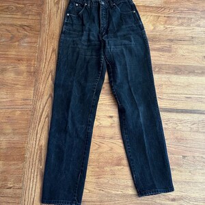 May include: A pair of black denim jeans with a classic five-pocket design. The jeans have a high waist and a straight leg cut. The dark wash denim shows subtle fading and wear, suggesting a vintage aesthetic. The jeans are laid flat on a wooden surface.