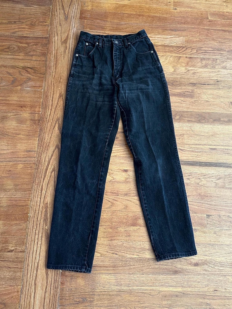 May include: A pair of black denim jeans with a classic five-pocket design. The jeans have a high waist and a straight leg cut. The dark wash denim shows subtle fading and wear, suggesting a vintage aesthetic. The jeans are laid flat on a wooden surface.