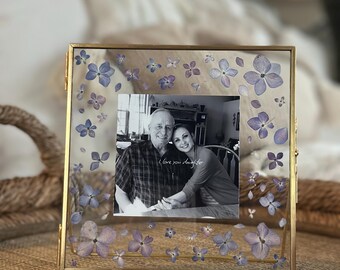 May include: A gold picture frame with a black and white photo of a man and woman. The frame is decorated with pressed purple flowers.