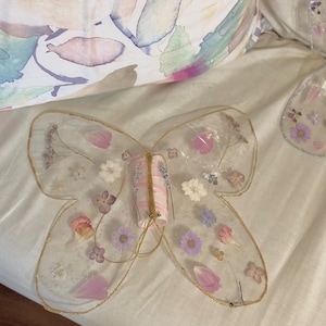 May include: A pair of clear resin butterfly wings with dried flowers and gold trim. The wings are attached to a pink and white striped fabric tube.