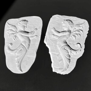Könnte beinhalten: Zwei weiße Gipsreliefskulpturen von Cherubinen. Jeder Cherub hat Flügel und ist in einer Tanzpose dargestellt. Die Skulpturen haben einen rauen, unregelmäßigen Rand und sind vor einem dunklen Hintergrund platziert.