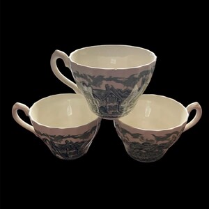 Może przedstawiać: Trzy białe filiżanki do herbaty z niebiesko-białym wzorem krajobrazu. Każda filiżanka ma zakrzywiony uchwyt i szeroką krawędź. Filiżanki ułożone są w trójkąt na czarnym tle.