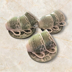 Może przedstawiać: Zestaw sześciu filiżanek i spodków. Filiżanki i spodki są białe z zielonym wzorem krajobrazu. Filiżanki mają zakrzywiony uchwyt, a spodki mają pasujący wzór obramowania. Zestaw jest ułożony na marmurowej powierzchni.