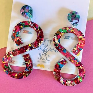 May include: A pair of colourful, abstract earrings. Each earring has a small, iridescent, circular stud connected to a larger, infinity-shaped design. The infinity shapes are filled with red, pink, and gold glitter. The earrings are displayed on a white card.