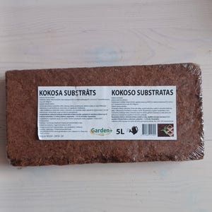Pode incluir: Um tijolo castanho de substrato de coco para jardinagem, etiquetado como "KOKOSO SUBSTRĀTS" em letão. O tijolo está envolto em plástico e tem uma etiqueta com o logotipo Garden+ e um ícone de regador. A etiqueta também indica "5L" e inclui um código de barras.