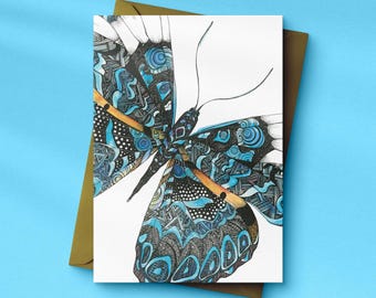 Könnte beinhalten: Eine Grußkarte mit einer detaillierten Illustration eines Schmetterlings. Der Schmetterling hat komplizierte Muster in Blau, Schwarz und Weiß. Das Design befindet sich auf einem weißen Hintergrund, mit einem goldenen Umschlag hinter der Karte. Die Karte liegt auf einer hellblauen Oberfläche.