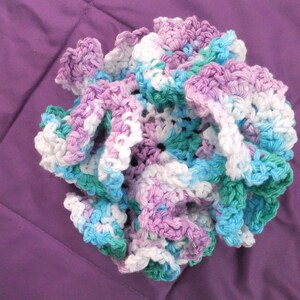 May include: A crocheted flower made with white, light blue, light purple, and green yarn. The flower is made with a ruffled design.