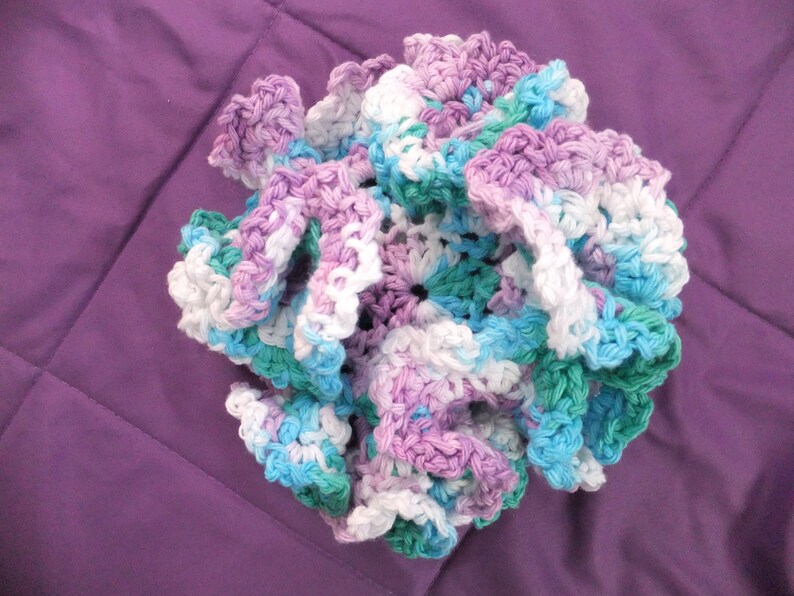May include: A crocheted flower made with white, light blue, light purple, and green yarn. The flower is made with a ruffled design.