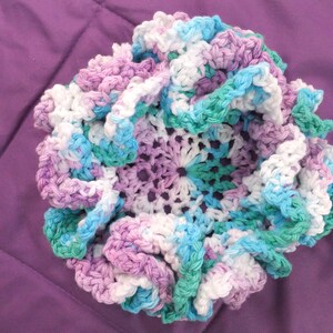 May include: A crocheted flower-shaped dish in shades of purple, blue, and white. The dish is made with a variety of stitches and has a ruffled edge.