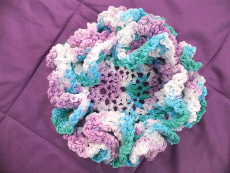 May include: A crocheted flower-shaped dish in shades of purple, blue, and white. The dish is made with a variety of stitches and has a ruffled edge.