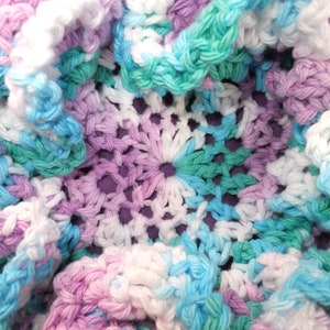 May include: A crocheted doily with a center design of white, light blue, and light green yarn. The doily is surrounded by a ruffled edge of white, light blue, and light purple yarn.