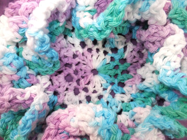 May include: A crocheted doily with a center design of white, light blue, and light green yarn. The doily is surrounded by a ruffled edge of white, light blue, and light purple yarn.