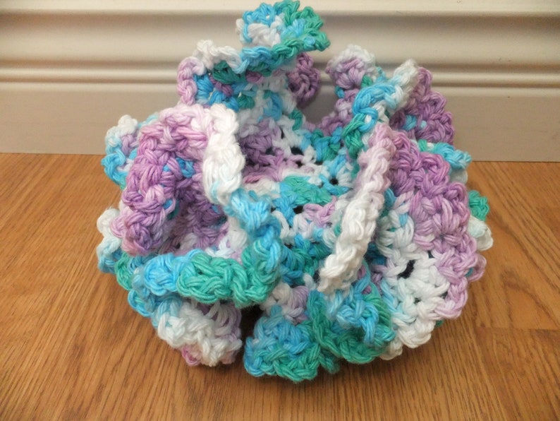 May include: A crocheted scrubby in shades of white, purple, blue, and green. The scrubby is made of soft yarn and has a ruffled texture.