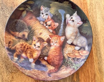 May include: A decorative ceramic plate featuring a whimsical scene of five kittens and a hedgehog. The kittens are depicted in various poses, with one standing on its hind legs, another peeking out from behind a tree, and others playing in the forest. The plate has a brown and green background with a birch tree trunk and mushrooms.