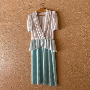 May include: A vintage dress with a white, wrap-style bodice and short sleeves. The dress features a light blue pleated skirt and a ruffled waist detail. The dress is hanging on a wooden hanger against a textured wall.