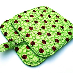 May include: Two square, quilted pot holders with a green background and a pattern of red ladybugs and white leaves. Each pot holder has a loop for hanging. The edges are trimmed with a green binding.