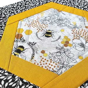 May include: A quilted fabric with a bee and floral design. The center features a white background with black and yellow bees, flowers, and honeycomb patterns. The center is framed by a yellow border, and the outer edge has a black and white leaf pattern.
