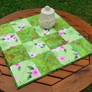 May include: A square quilted table topper featuring a patchwork design of light green fabric with pink floral patterns and dark green marbled textures, topped with a cream-colored ceramic jar decorated with embossed seashells and a starfish.