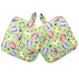 May include: Two square green pot holders with a colorful Easter egg pattern. The eggs are decorated with stripes, polka dots, and floral designs. Each pot holder has a matching fabric loop for hanging.