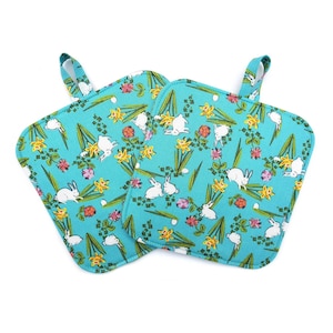 May include: Two square turquoise pot holders with a loop for hanging. The fabric features a pattern of white rabbits, yellow daffodils, and pink and orange flowers. The pot holders are designed for kitchen use.