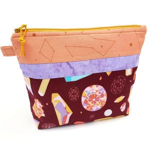 May include: A zippered pouch featuring a top section of peach fabric with thin constellation lines, a middle band of mottled light purple fabric, and a bottom section of dark maroon fabric patterned with colorful geometric crystal illustrations.