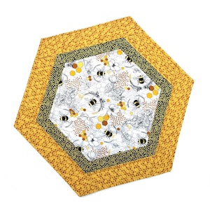 May include: A hexagonal quilted placemat with a bee theme. The outer layer is yellow with a honeycomb pattern. The middle layer is black with white dots. The center layer is white with bees, flowers, and honeycomb designs.