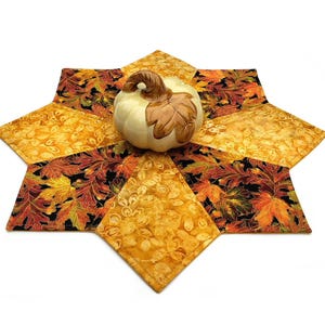 May include: A decorative fall-themed centerpiece featuring a cream-colored pumpkin with a brown leaf accent. The pumpkin sits atop a star-shaped quilted placemat with a leaf pattern in shades of orange, yellow, and brown.
