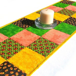 May include: A rectangular patchwork table runner featuring a grid of squares in orange, yellow, lime green, and black. The black squares display small pumpkin and bat patterns, while other squares feature swirl designs. A white pillar candle sits on top.