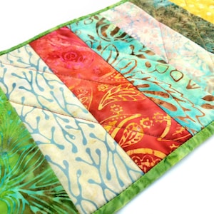 May include: A colorful, quilted placemat with a green border. The design features diagonal stripes in various patterns and colors, including green, red, blue, and yellow. The fabrics have floral and abstract designs.