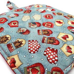 May include: A square fabric potholder with a light blue background featuring a repeating pattern of various oven mitts in red, white, and tan colors, accented with small white dots and a red fabric border with a small hanging loop.