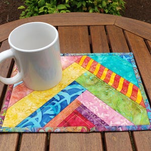 May include: A white ceramic mug rests on a colorful, quilted coaster. The coaster features a patchwork design with diagonal stripes in shades of yellow, blue, red, green, and pink. The mug is empty and sits on a wooden table.