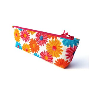 May include: A rectangular pencil case featuring a white background with a repeating pattern of vibrant daisy flowers in shades of orange, yellow, pink, and blue. The case is secured with a bright pink zipper closure along the top edge.