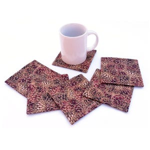 May include: A set of six square coasters featuring a repeating pattern of brown and tan pinecones, with one plain white ceramic mug resting on top of one coaster. The coasters have stitched edges and a flat, fabric-like surface texture.