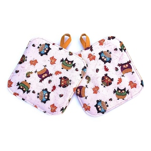 May include: Two square pot holders featuring a light pink background with a repeating pattern of colorful owls, orange and red autumn leaves, and small brown acorns. Each pot holder includes a small mustard yellow fabric hanging loop at the top.