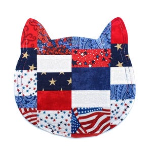 May include: A patriotic cat-shaped quilted coaster featuring a patchwork design. The coaster incorporates red, white, and blue fabrics with star and flag patterns. The cat's ears are solid red, and the overall shape is approximately 7 inches wide.