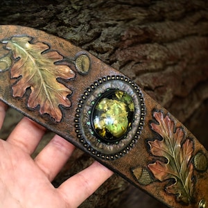 May include: A brown leather cuff bracelet with embossed oak leaves and acorns. The bracelet features a central oval-shaped embellishment with a green and gold design, surrounded by a beaded border. The leaves are detailed with shades of brown and green.