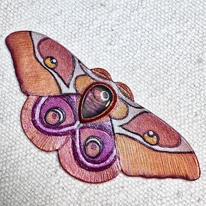 以下が含まれることがあります： 精巧なディテールが施された、手作りの蛾の形をしたブローチ。ブローチはオレンジ、ピンク、紫、ゴールドの色使いです。中央には涙型のイリデセントストーンがセットされています。羽はテクスチャード加工されています。