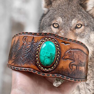 Peut inclure: Bracelet manchette en cuir marron avec une grande pierre ovale en malachite. Le bracelet présente des motifs de forêt et d'ours en relief, avec des clous en laiton et des coutures vertes. Un loup est en arrière-plan.