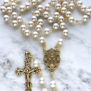 May include: A rosary featuring lustrous white faux pearl beads linked by gold-tone metal wire. The piece includes a gold-tone metal centerpiece depicting a figure and a detailed gold-tone crucifix with an ornate design and the letters INRI at the top.