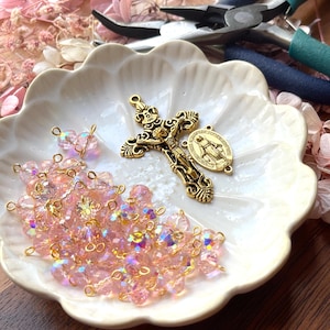 May include: A white, scalloped dish holds a gold-colored crucifix, a religious medal, and strands of pink crystal beads. Pliers with blue and black handles are in the background. The beads and crucifix suggest a theme of religious jewelry making.