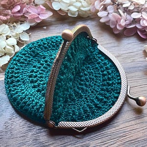 May include: A handmade, teal green crochet coin purse with a vintage-style metal clasp. The purse is round and open, revealing the textured crochet stitches. The clasp is a bronze color, and the purse is set against a wooden surface with dried flowers.