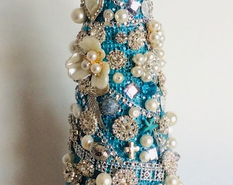 Puede incluir: Un adorno de árbol de Navidad de forma cónica de color azul turquesa cubierto de pedrería, perlas y otros adornos decorativos. La parte superior del árbol está adornada con una estrella de cristal transparente.
