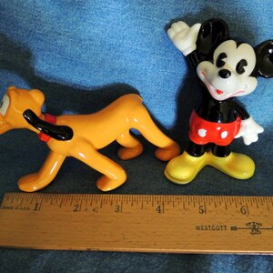 May include: A ceramic figurine of Mickey Mouse wearing red shorts with white polka dots, standing with his right arm raised, and a ceramic figurine of Pluto, a yellow dog with black ears, running. Both figurines are approximately 5 inches tall.