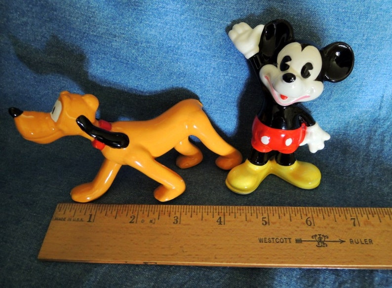 May include: A ceramic figurine of Mickey Mouse wearing red shorts with white polka dots, standing with his right arm raised, and a ceramic figurine of Pluto, a yellow dog with black ears, running. Both figurines are approximately 5 inches tall.