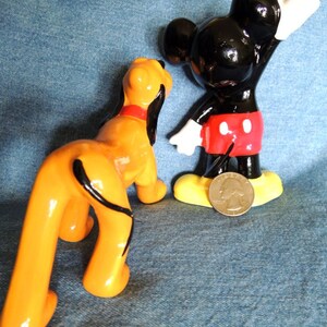 May include: A ceramic figurine of Mickey Mouse wearing his signature red shorts and yellow shoes, standing with his right arm raised.  A ceramic figurine of Pluto, Mickey's dog, is standing next to him.  Pluto is orange and black with a long tail.