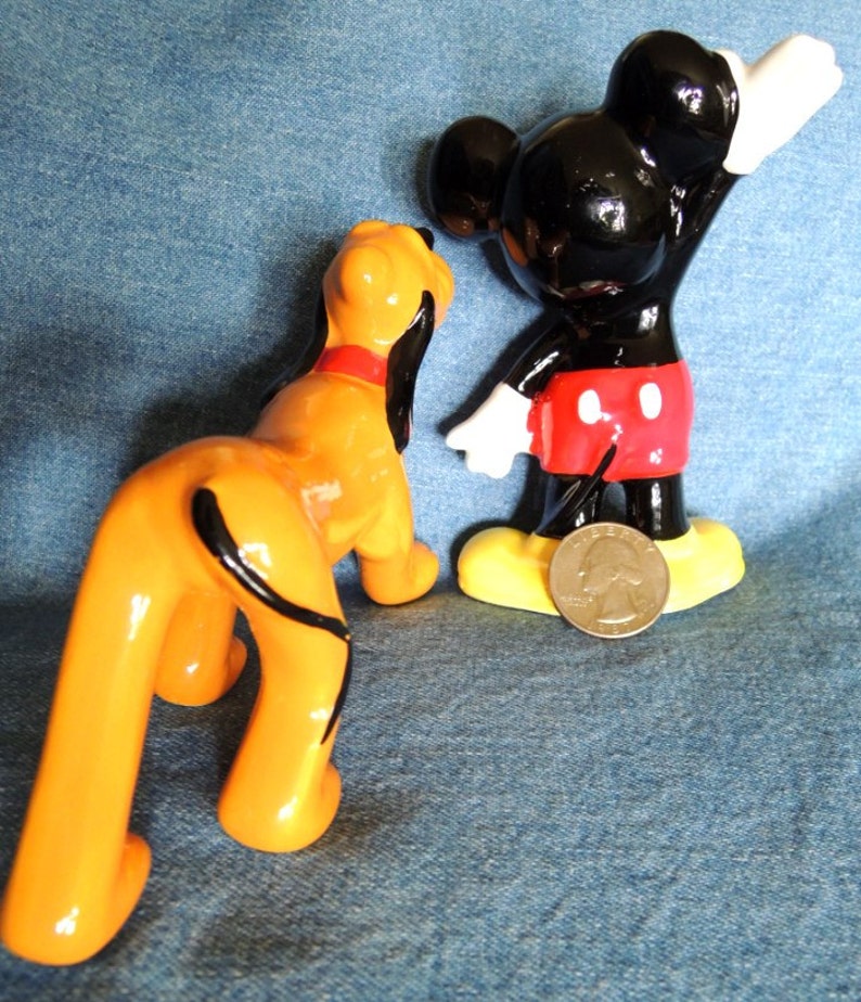 May include: A ceramic figurine of Mickey Mouse wearing his signature red shorts and yellow shoes, standing with his right arm raised.  A ceramic figurine of Pluto, Mickey's dog, is standing next to him.  Pluto is orange and black with a long tail.