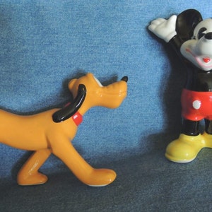 May include: A ceramic figurine of Pluto the dog, in a yellow-orange color, with black ears and a red collar, is standing on a blue denim surface.  A ceramic figurine of Mickey Mouse, in black with red shorts and yellow shoes, is standing on the same surface, waving with his right hand.