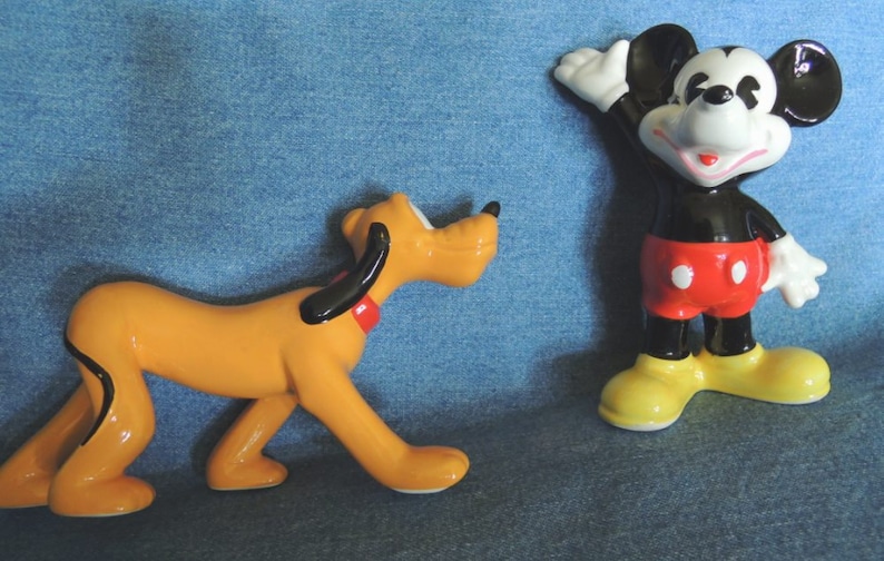 May include: A ceramic figurine of Pluto the dog, in a yellow-orange color, with black ears and a red collar, is standing on a blue denim surface.  A ceramic figurine of Mickey Mouse, in black with red shorts and yellow shoes, is standing on the same surface, waving with his right hand.