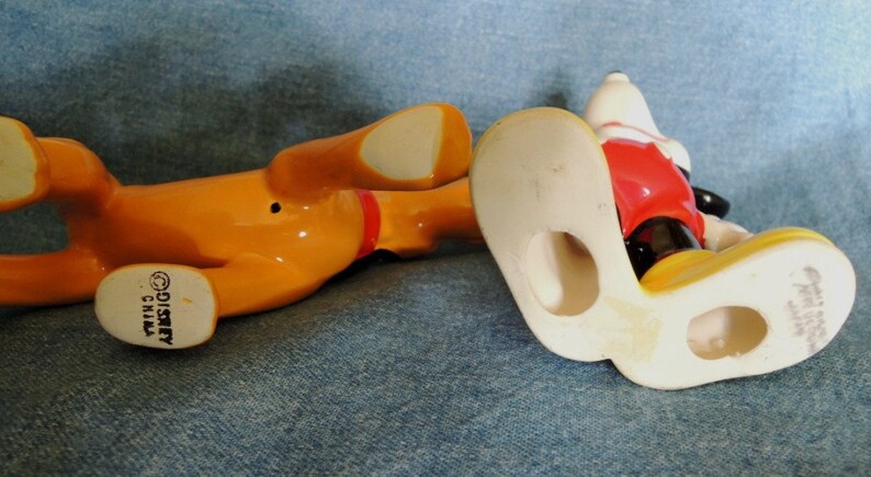 May include: Bottom view of two ceramic figurines, one of Mickey Mouse and one of Pluto. Mickey Mouse is wearing red shorts and yellow shoes. Pluto is orange and has the text "&copy;DISNEY CHINA" on the bottom.